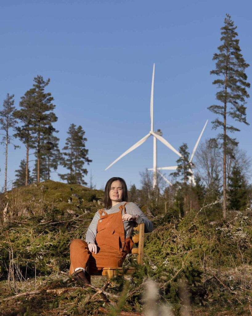 Porträttbild av Martha Kristensen sittandes på en stol framför tre vindkraftverk, stående format
