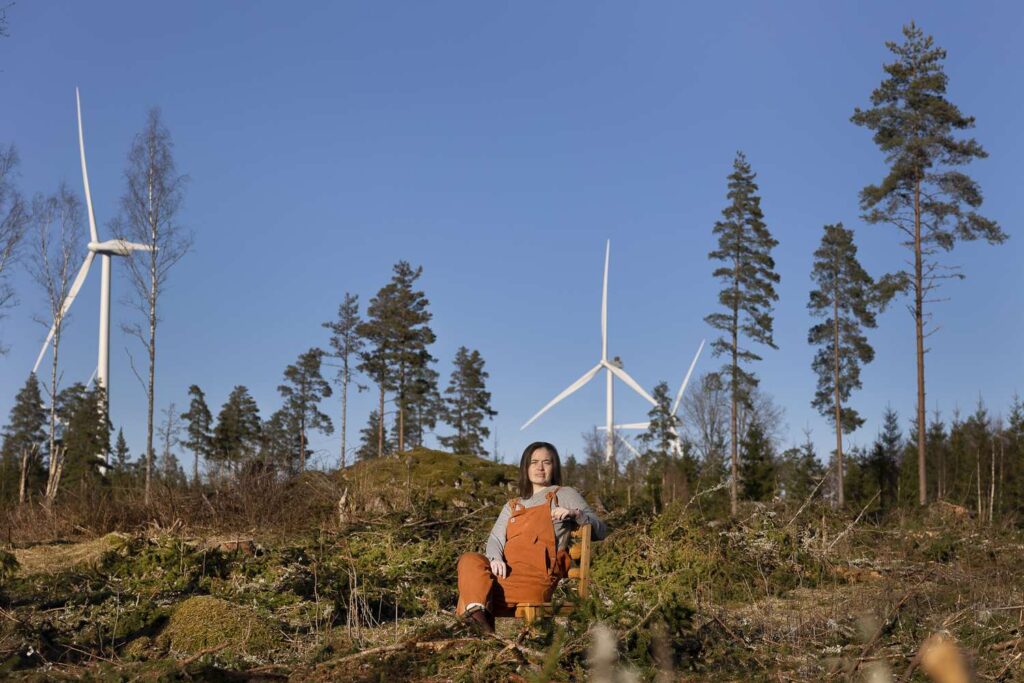 Porträttbild av Martha Kristensen sittandes på en stol framför tre vindkraftverk, liggande format